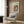 May include: Framed Renoir reproduction poster showing mother holding child in soft pastels, displayed on wood panel wall above cream upholstered armchair with geometric throw pillow. Modern brass floor lamp and dark side table with open book complete the warm, contemporary living space scene.