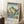May include: Framed Japanese woodblock print showing dramatic coastal landscape with rocky cliffs, waterfall, and blue water. Traditional ukiyo-e art style with green hills, yellow rock formations, and cherry blossoms. Print features Japanese text and English caption reading "Noto Province, Waterfall Bay." Displayed in light wood frame leaning against white wall next to small wooden table with white coffee cup and beige throw blanket on textured white rug.
