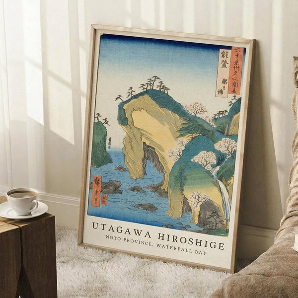 May include: Framed Japanese woodblock print showing dramatic coastal landscape with rocky cliffs, waterfall, and blue water. Traditional ukiyo-e art style with green hills, yellow rock formations, and cherry blossoms. Print features Japanese text and English caption reading "Noto Province, Waterfall Bay." Displayed in light wood frame leaning against white wall next to small wooden table with white coffee cup and beige throw blanket on textured white rug.