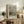 May include: Van Gogh museum poster featuring fishing boats on a beach in muted tones, displayed in light wood frame on rustic wooden sideboard. Room styled with fern plant in white pot, cream upholstered chair with brown leather cushion, stacked books, and ceramic vase. Soft natural lighting creates warm, earthy atmosphere with beige walls.