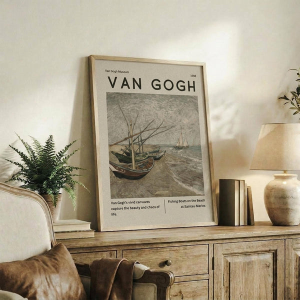 May include: Van Gogh museum poster featuring fishing boats on a beach in muted tones, displayed in light wood frame on rustic wooden sideboard. Room styled with fern plant in white pot, cream upholstered chair with brown leather cushion, stacked books, and ceramic vase. Soft natural lighting creates warm, earthy atmosphere with beige walls.