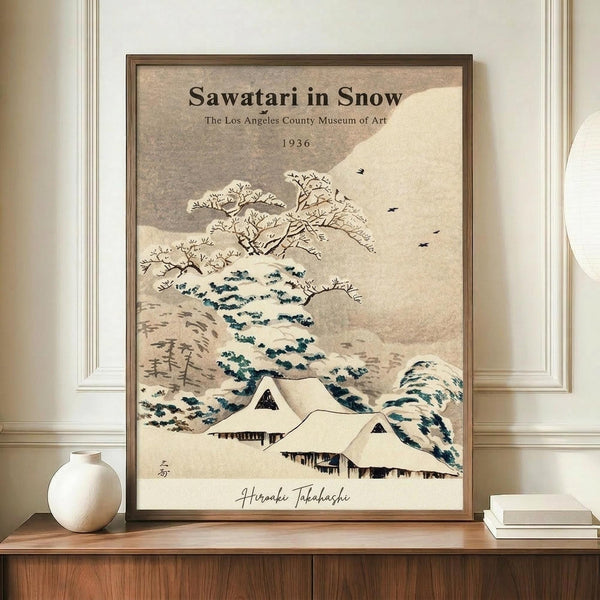 May include: Framed vintage Japanese woodblock print showing snow-covered houses with steep roofs, bare winter trees, and rolling hills in muted beige and blue tones. The artwork sits on a wooden sideboard between a round white ceramic vase and stacked books against cream wainscoted walls. Museum exhibition text and artist signature visible on the print.