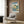 May include: Japanese woodblock print showing coastal landscape with green hills, rock formations, and blue water displayed in light wood frame on textured wall above beige upholstered chair with geometric patterned pillow. Modern brass floor lamp and dark side table with open book complete the neutral-toned room scene.