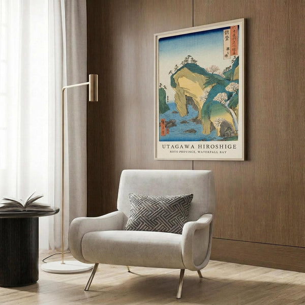 May include: Japanese woodblock print showing coastal landscape with green hills, rock formations, and blue water displayed in light wood frame on textured wall above beige upholstered chair with geometric patterned pillow. Modern brass floor lamp and dark side table with open book complete the neutral-toned room scene.