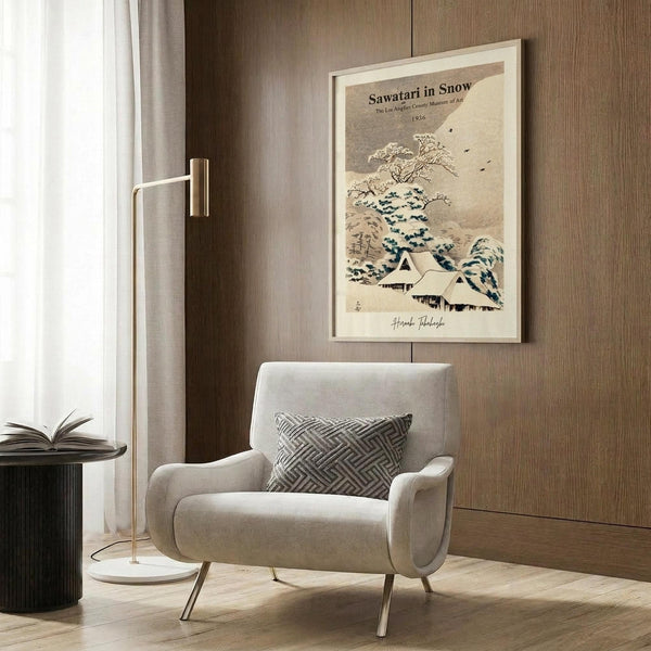 May include: Framed vintage-style Japanese winter landscape poster showing snow-covered houses and bare tree with blue and beige tones, displayed on wood-paneled wall above beige upholstered armchair with geometric patterned pillow, beside brass floor lamp and black side table with open book