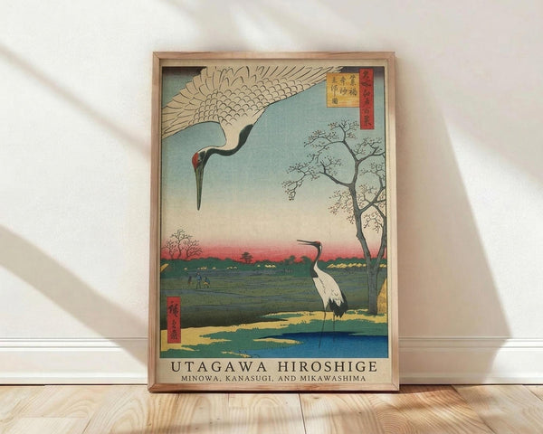 May include: Framed Japanese woodblock print featuring two cranes in a serene landscape with traditional ukiyo-e styling. One crane flies overhead with outstretched wings while another stands by a blue stream. Scene shows bare winter trees, distant pagoda, and gradient sky from blue to pink. Artwork displays authentic aged colors and Japanese text cartouches. Natural wood frame sits on light wooden floor against white wall with soft natural lighting.