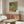 May include: Japanese art poster featuring figures and a tiger on green patterned background, displayed in thin frame above wooden sideboard. Modern living room setting with white curved chair, glass oval coffee table, and minimalist floor lamp. Warm natural lighting creates shadows on white walls.