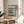 May include: Framed Japanese woodblock print featuring a coastal scene with turquoise waters, golden rocky cliffs, small boats, and Mount Fuji in the distance. The artwork displays traditional ukiyo-e style with red text cartouches and sits on a round wooden side table next to a beige upholstered chair. Sunlight streams across white walls creating geometric shadows, with green plant leaves visible in the corner.