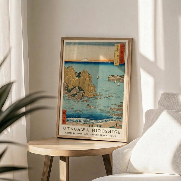 May include: Framed Japanese woodblock print featuring a coastal scene with turquoise waters, golden rocky cliffs, small boats, and Mount Fuji in the distance. The artwork displays traditional ukiyo-e style with red text cartouches and sits on a round wooden side table next to a beige upholstered chair. Sunlight streams across white walls creating geometric shadows, with green plant leaves visible in the corner.