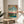 May include: Japanese woodblock print-style artwork in wooden frame showing two cranes - one flying with outstretched wings above, one standing below near water. Turquoise sky transitions to pink sunset horizon with bare tree silhouettes. Print sits on round wooden side table next to cream armchair, with person's hand reaching toward frame. Soft natural lighting creates shadows on white wall. Green plant leaves visible in corner.