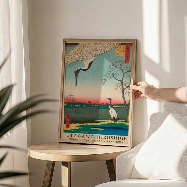 May include: Japanese woodblock print-style artwork in wooden frame showing two cranes - one flying with outstretched wings above, one standing below near water. Turquoise sky transitions to pink sunset horizon with bare tree silhouettes. Print sits on round wooden side table next to cream armchair, with person's hand reaching toward frame. Soft natural lighting creates shadows on white wall. Green plant leaves visible in corner.