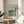 May include: A framed Van Gogh museum poster featuring a swirling landscape painting with rolling green hills, dramatic cloudy sky in blues and whites, and golden wheat fields. The poster sits on a round wooden side table next to a beige upholstered chair with a throw pillow. Natural sunlight streams across the neutral-toned room, casting soft shadows. A plant's leaves are partially visible in the corner.