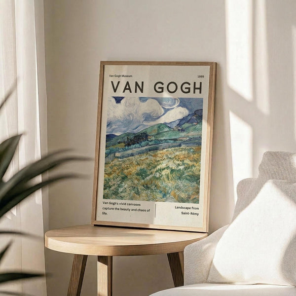 May include: A framed Van Gogh museum poster featuring a swirling landscape painting with rolling green hills, dramatic cloudy sky in blues and whites, and golden wheat fields. The poster sits on a round wooden side table next to a beige upholstered chair with a throw pillow. Natural sunlight streams across the neutral-toned room, casting soft shadows. A plant's leaves are partially visible in the corner.