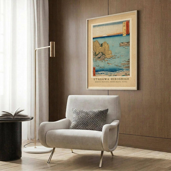 May include: Japanese woodblock print artwork displayed in modern living room with beige armchair, geometric pillow, brass floor lamp, and dark wood paneling. The framed poster shows a coastal scene with rocky outcrops, blue ocean waters, and traditional Japanese artistic style in muted blues, browns, and coral tones.
