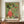 May include: A framed Japanese woodblock print reproduction displayed on a wooden sideboard against white wainscoting. The artwork features a monk in red robes seated on a tiger, surrounded by various animals and figures on a green geometric patterned background. A small white ceramic vase and stacked books sit beside the brown-framed print, which shows rich colors including orange, green, and coral tones in the traditional ukiyo-e style.