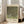 May include: Framed art print leaning against white wall next to beige sofa in bright room with large windows. Print shows Klimt's impressionistic painting of green foliage and rosebushes in dappled light with cream colored border text. Natural wood frame, approximately 2-3 feet tall, positioned on light wooden floor with partial view of area rug.
