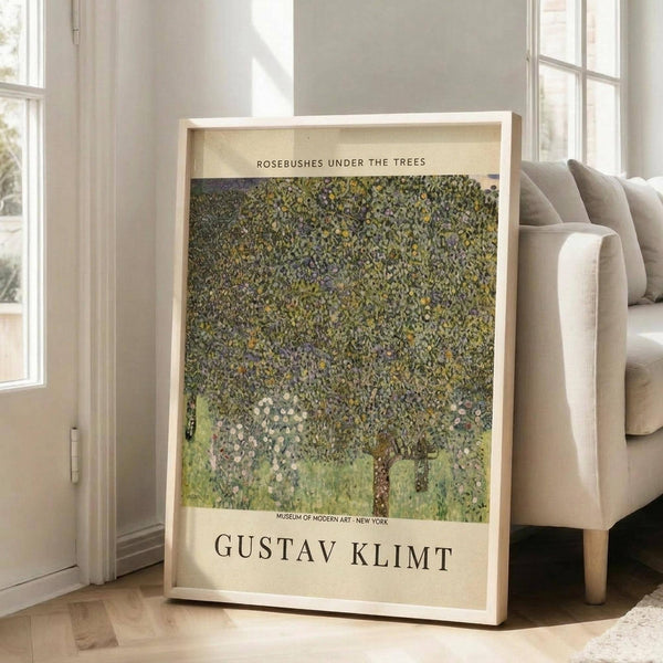 May include: Framed art print leaning against white wall next to beige sofa in bright room with large windows. Print shows Klimt's impressionistic painting of green foliage and rosebushes in dappled light with cream colored border text. Natural wood frame, approximately 2-3 feet tall, positioned on light wooden floor with partial view of area rug.