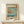 May include: Framed Japanese woodblock print featuring coastal scene with golden cliffs, turquoise water, small boats, and Mount Fuji in distance. Print displays in light wood frame on white wooden surface beside small white wrapped gift with greenery. Cream wall background with natural lighting creates shadows.