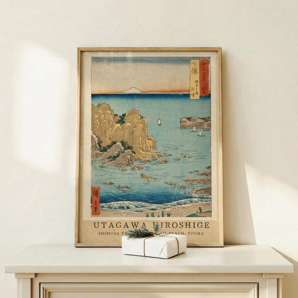 May include: Framed Japanese woodblock print featuring coastal scene with golden cliffs, turquoise water, small boats, and Mount Fuji in distance. Print displays in light wood frame on white wooden surface beside small white wrapped gift with greenery. Cream wall background with natural lighting creates shadows.