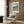 May include: Beige upholstered armchair with geometric patterned pillow positioned next to wood-paneled wall. Framed Van Gogh landscape poster featuring swirling clouds over green fields hangs above chair. Brass floor lamp and dark round side table with open book visible. Natural light from window with white curtains illuminates warm-toned modern interior space with hardwood flooring.