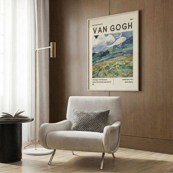May include: Beige upholstered armchair with geometric patterned pillow positioned next to wood-paneled wall. Framed Van Gogh landscape poster featuring swirling clouds over green fields hangs above chair. Brass floor lamp and dark round side table with open book visible. Natural light from window with white curtains illuminates warm-toned modern interior space with hardwood flooring.