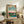 May include: Framed Japanese woodblock print featuring two cranes in a landscape with blue water, red sunset sky, and bare tree, displayed on rustic wooden sideboard alongside fern plant, books, and ceramic vase in neutral-toned living room setting