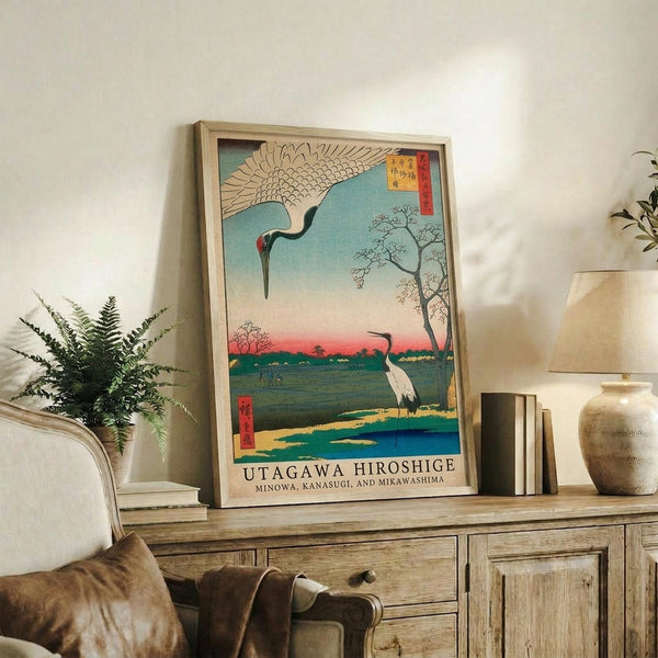 May include: Framed Japanese woodblock print featuring two cranes in a landscape with blue water, red sunset sky, and bare tree, displayed on rustic wooden sideboard alongside fern plant, books, and ceramic vase in neutral-toned living room setting