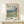 May include: Framed Van Gogh landscape poster showing swirling clouds over rolling hills and grasslands in blues, greens, and golden yellows. The print features museum-style text and is displayed in a natural wood frame on a light wooden surface next to a small wrapped gift with twine and evergreen sprig against a cream wall.