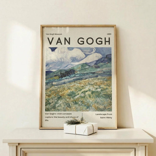 May include: Framed Van Gogh landscape poster showing swirling clouds over rolling hills and grasslands in blues, greens, and golden yellows. The print features museum-style text and is displayed in a natural wood frame on a light wooden surface next to a small wrapped gift with twine and evergreen sprig against a cream wall.