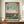 May include: Japanese woodblock print artwork displayed in brown frame on wooden credenza beside white ceramic vase and books. The vintage-style print features two cranes - one flying overhead with outstretched wings, one standing by blue water - against sunset sky with bare tree branches. Traditional red Japanese text cartouches visible in corners. Cream colored walls with white trim molding in background.