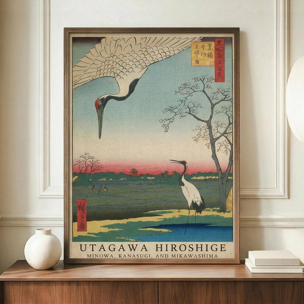 May include: Japanese woodblock print artwork displayed in brown frame on wooden credenza beside white ceramic vase and books. The vintage-style print features two cranes - one flying overhead with outstretched wings, one standing by blue water - against sunset sky with bare tree branches. Traditional red Japanese text cartouches visible in corners. Cream colored walls with white trim molding in background.