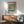 May include: Japanese woodblock print artwork displayed in wooden frame on white wall above mid-century modern wooden sideboard. Green upholstered chair with wooden arms sits nearby. Print shows coastal scene with golden rock formations, blue water, small boats, and Mount Fuji in distance. Warm natural lighting creates shadows across minimalist room setting.