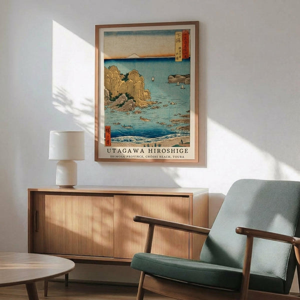 May include: Japanese woodblock print artwork displayed in wooden frame on white wall above mid-century modern wooden sideboard. Green upholstered chair with wooden arms sits nearby. Print shows coastal scene with golden rock formations, blue water, small boats, and Mount Fuji in distance. Warm natural lighting creates shadows across minimalist room setting.
