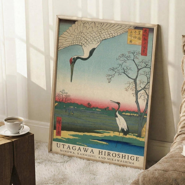 May include: Framed Japanese woodblock print featuring two cranes in a pastoral landscape with flowering tree, mountains, and traditional text cartouches. The artwork shows muted blues, greens, and coral tones in classic ukiyo-e style. Print leans against white wall on white textured rug next to wooden side table with coffee cup and cozy throw blanket in minimalist room setting.