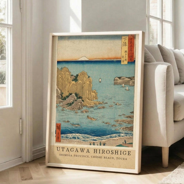 May include: Framed Japanese woodblock print featuring a coastal scene with turquoise waters, rocky cliffs, small boats, and Mount Fuji in the distance. The artwork displays traditional ukiyo-e style with muted earth tones and blue hues, positioned on light wooden floors next to a beige sofa in a bright room with white walls and large windows.