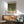 May include: A framed art print featuring Gustav Klimt's "Rosebushes Under the Trees" displayed above a mid-century modern wood credenza. The artwork shows a dense canopy of green foliage with white flowers beneath in Klimt's distinctive style. A sage green upholstered armchair with wooden frame sits beside the credenza, with a white ceramic table lamp and round wooden coffee table visible. Warm sunlight streams across the white wall, creating geometric shadows in this contemporary living