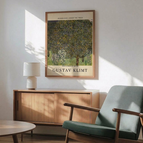 May include: A framed art print featuring Gustav Klimt's "Rosebushes Under the Trees" displayed above a mid-century modern wood credenza. The artwork shows a dense canopy of green foliage with white flowers beneath in Klimt's distinctive style. A sage green upholstered armchair with wooden frame sits beside the credenza, with a white ceramic table lamp and round wooden coffee table visible. Warm sunlight streams across the white wall, creating geometric shadows in this contemporary living