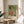 May include: Framed Japanese woodblock print showing figures with a tiger on green geometric background, displayed on light wood round side table next to beige upholstered chair. Sunlight streams across white wall creating soft shadows. Green plant leaves visible in corner. Print features traditional art style with monk in red robes, tiger with black stripes, and various other figures in colorful clothing against honeycomb pattern backdrop.