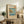 May include: Framed Japanese woodblock print featuring coastal scene with rocky cliffs, blue ocean waters, and Mount Fuji in distance, displayed on rustic wooden sideboard alongside fern plant, ceramic vase, and stacked books in neutral-toned living room setting with beige armchair visible.