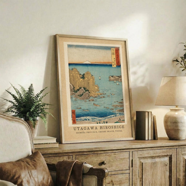 May include: Framed Japanese woodblock print featuring coastal scene with rocky cliffs, blue ocean waters, and Mount Fuji in distance, displayed on rustic wooden sideboard alongside fern plant, ceramic vase, and stacked books in neutral-toned living room setting with beige armchair visible.