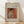 May include: Framed art print leaning against white wall on wooden floor with natural sunlight casting shadows. Features Renoir's 1908 painting of woman in white dress sewing, set against warm orange-red background. Print has cream matting with elegant script text identifying the artwork and artist. Thin wooden frame in natural wood tone complements the soft, impressionist brushwork and peachy color palette of the classic painting.