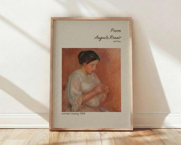 May include: Framed art print leaning against white wall on wooden floor with natural sunlight casting shadows. Features Renoir's 1908 painting of woman in white dress sewing, set against warm orange-red background. Print has cream matting with elegant script text identifying the artwork and artist. Thin wooden frame in natural wood tone complements the soft, impressionist brushwork and peachy color palette of the classic painting.