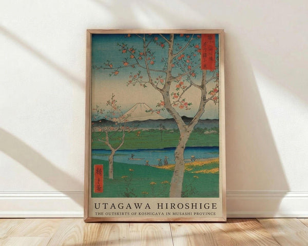 May include: A framed Japanese woodblock print reproduction leaning against a white wall on light wood flooring. The artwork depicts a spring landscape with blooming cherry trees in the foreground, Mount Fuji in the distance, and a blue river winding through green fields. Small figures are visible along the riverbank. The print features traditional ukiyo-e style with vibrant blues, greens, and coral pink blossoms. Red Japanese text cartouches appear in the upper right and lower left corners. The