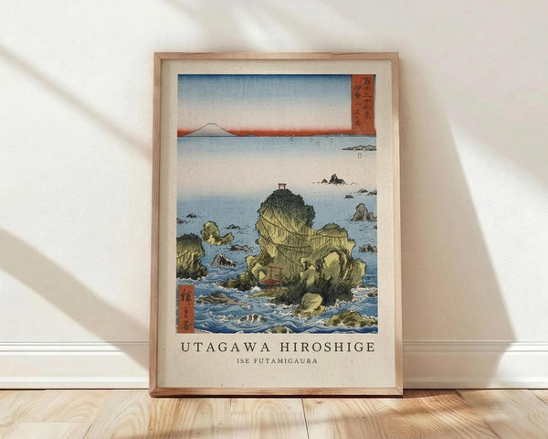 May include: Framed Japanese woodblock print reproduction showing coastal rock formations with a small torii gate, surrounded by blue ocean waters and distant mountains. The artwork features traditional ukiyo-e style with soft blues, greens, and earth tones, displayed in a light wood frame against a white wall on wooden flooring with natural window light casting shadows.
