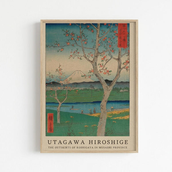 Framed Japanese woodblock print showing a blossoming tree in the foreground with pink flowers, overlooking a scenic landscape with Mount Fuji in the distance. The artwork features traditional ukiyo-e style with blue sky, green fields, a winding river, and small figures near the water. Red text cartouches appear in the upper right and lower left corners. The print is displayed in a light wooden frame against a white wall, with the artist name "Utagawa Hiroshige" and artwork title printed below th