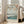 May include: A framed Japanese woodblock print displaying cherry blossoms over Mount Fuji and a river landscape in soft blues, greens, and coral tones. The artwork features traditional Japanese calligraphy and shows boats on water with trees along the shoreline. The light wood frame leans against a white wall beside a beige upholstered chair in a bright, minimalist room with natural sunlight streaming through tall windows onto hardwood floors.