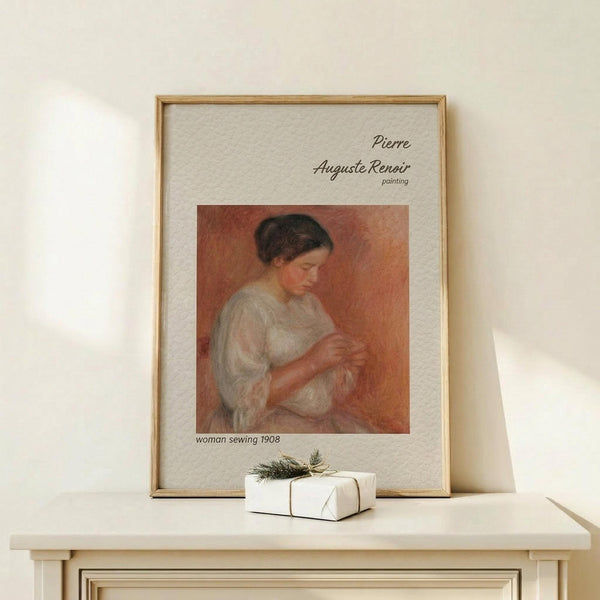 May include: Framed art print of Pierre Auguste Renoir's "Woman Sewing" 1908 painting showing a woman in white garment concentrating on needlework against warm orange background. Natural wood frame with cream matting displays the classic impressionist artwork. Print sits on light wooden surface next to small white wrapped gift with green sprig and twine, against neutral beige wall.