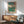 May include: Japanese woodblock print poster in wooden frame hanging on white wall above mid-century modern wood sideboard with white ceramic lamp. Green upholstered armchair with wooden arms sits nearby. Print shows cherry blossom tree overlooking blue-green landscape with Mount Fuji in distance. Warm sunlight creates shadows across the minimalist room with wooden floors.