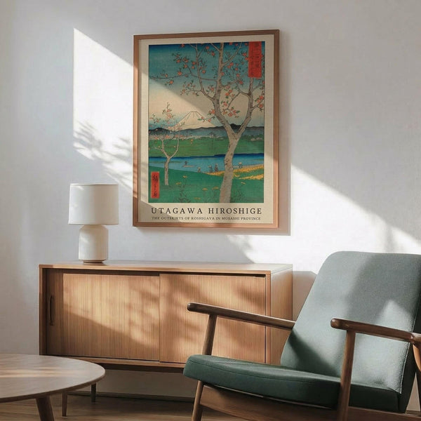 May include: Japanese woodblock print poster in wooden frame hanging on white wall above mid-century modern wood sideboard with white ceramic lamp. Green upholstered armchair with wooden arms sits nearby. Print shows cherry blossom tree overlooking blue-green landscape with Mount Fuji in distance. Warm sunlight creates shadows across the minimalist room with wooden floors.