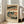 May include: Framed Japanese woodblock print showing a scenic landscape with buildings, bridge, and small figures in traditional dress. Print features muted tones of teal, coral, and cream with Japanese text at top. Light wood frame leans against white wall next to beige upholstered sofa in bright, modern living room with large windows and natural light casting shadows on hardwood floors.